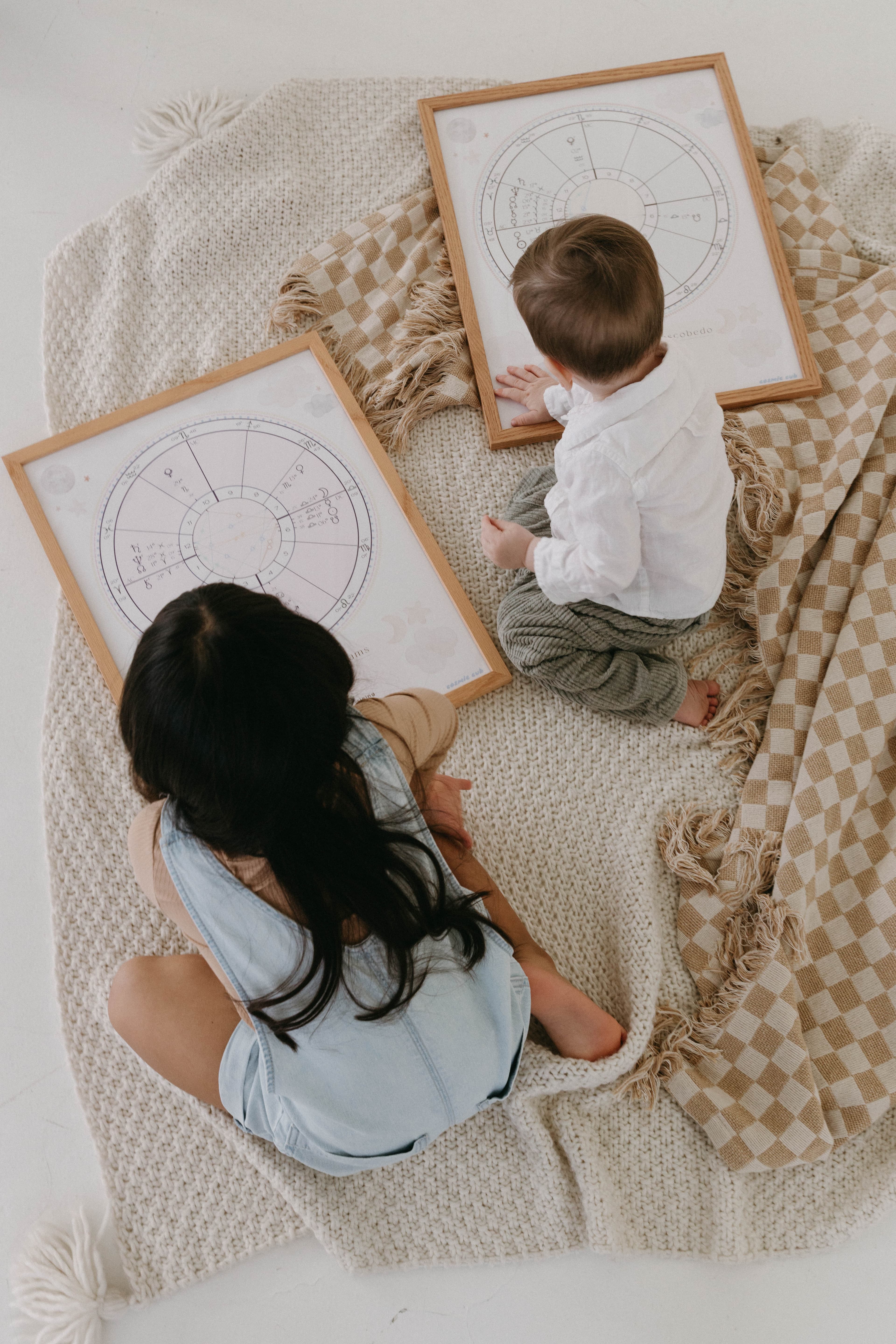 A mother and baby exploring two framed natal chart portraits laid on a knit blanket